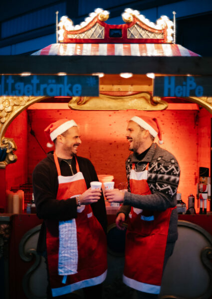 Zwei Männer mit Weihnachtsmützen stoßen auf einem Weihnachtsmarkt lachend mit Glühwein an