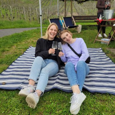 Zwei Frauen sitzen in den Weinbergen auf einer Picknickdecke und prosten sich zu