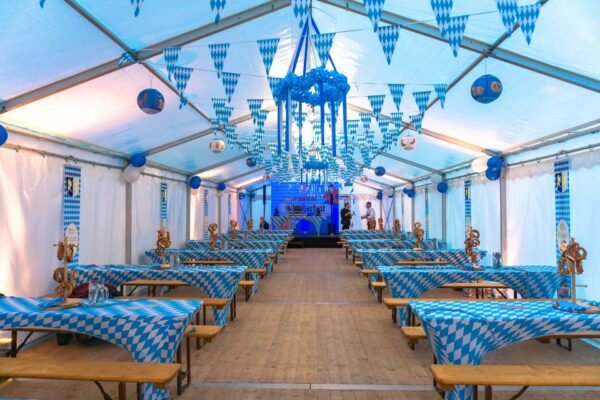 Dekoriertes Festzelt mit blau-weißen Girlanden, Tischdecken, Luftballons, Brezeln und kleiner Bühne
