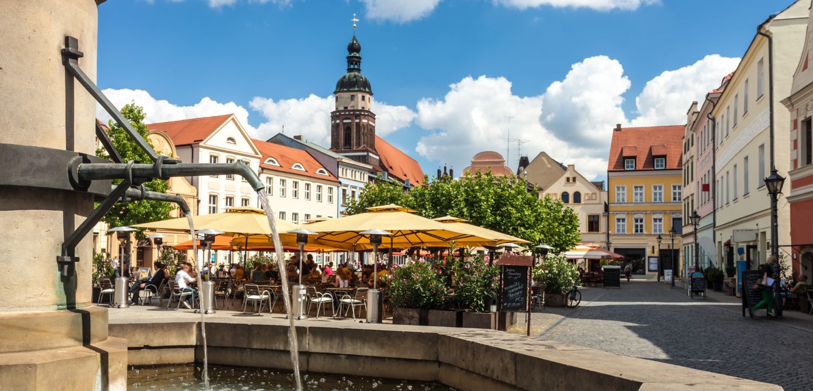 Brunnen im Vordergrund, Gastronomie mit Schirmen, Häuserfronten und Kirchturm im Hintergrund