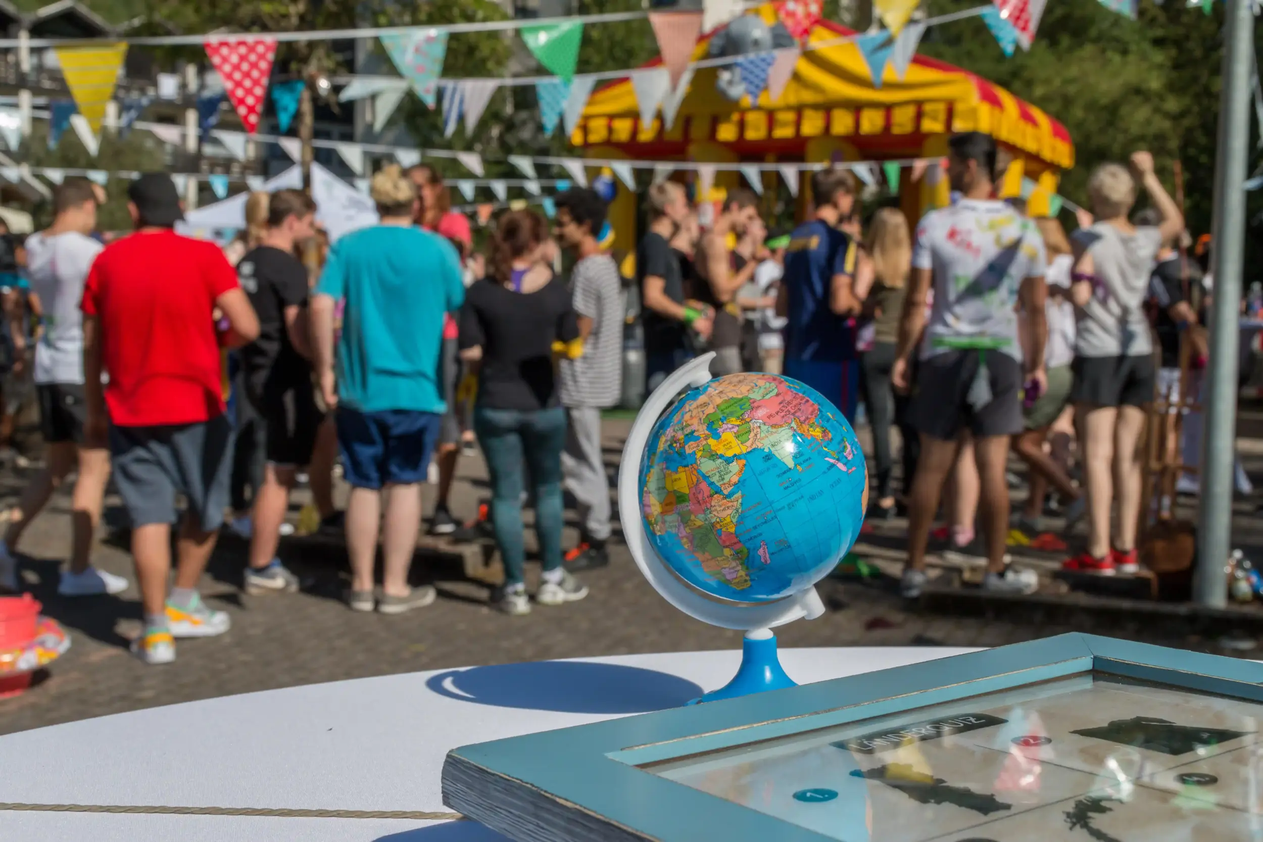 Auf einem Tisch steht ein Globus, im Hintergrund ist eine Menschenmenge, sowie dekorierte Pavillions