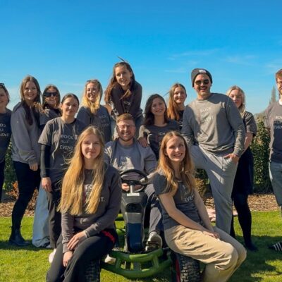 Vierzehn Personen lächeln in die Kamera, im Hintergrund ist grünes Gras und blauer Himmel