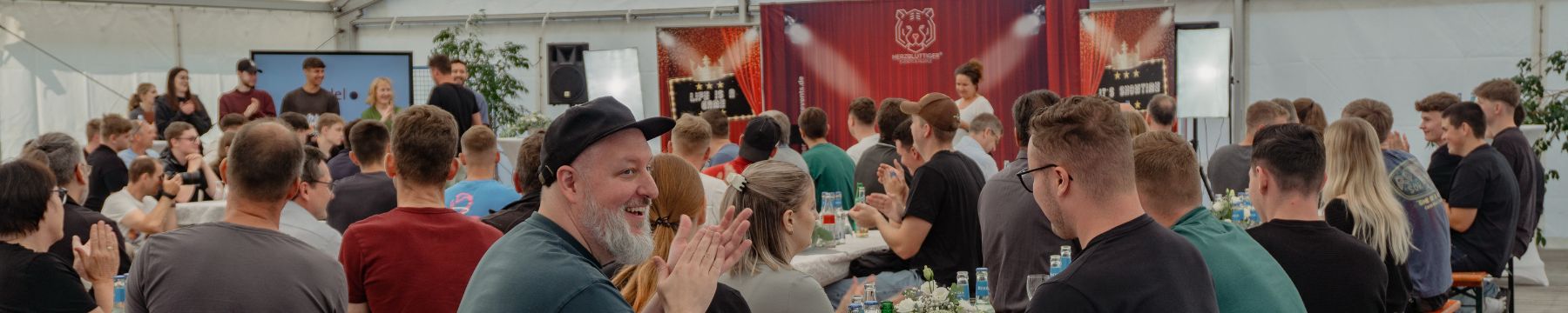 Personen sitzen an Biertischen, ein Mann lächelt in Richtung der Kamera, im Hintergrund ist eine Showbühne aufgebaut