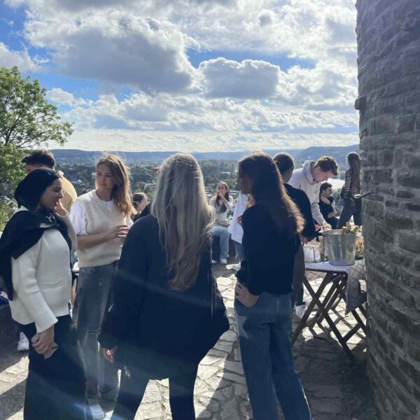 Mehrere Personen stehen auf einer steinernen Aussichtsterrasse und unterhalten sich. Im Hintergrund erstreckt sich ein weiter Blick über eine Stadtlandschaft und Hügel unter dem Himmel. Rechts steht ein Tisch mit Getränken neben einer Steinmauer.
