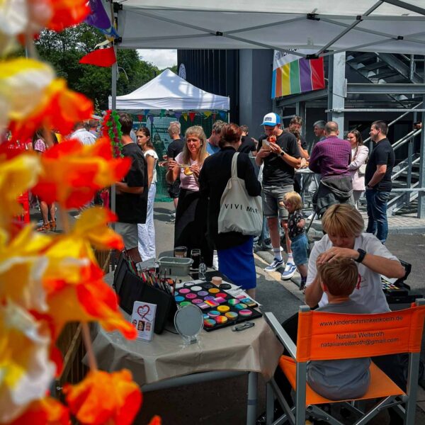 Menschen besuchen ein Straßenfest mit Zelten und Regenbogenflagge, während an einem Tisch ein Kind geschminkt wird.