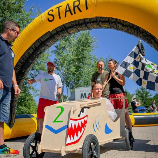 Menschen stehen unter einem gelben Startbogen, während eine Frau in einem selbstgebauten Holz-Seifenkistenauto mit Haifischmotiv sitzt und zum Rennen bereit ist.