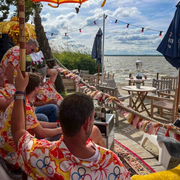 Mehrere Menschen in bunten Hemden sitzen an einer dekorierten Strandterrasse am Wasser, während ein großer Pokal auf einem Tisch mit Blick auf den See steht.